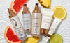 Five skincare products laying in water with pieces of grapefruit, pineapple and lemon slices. 